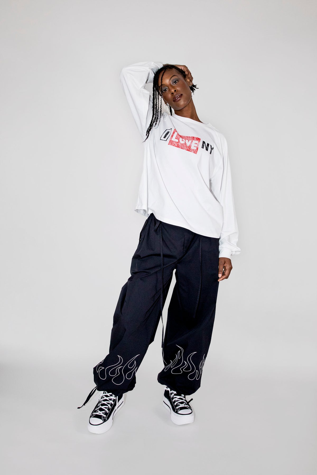 A model posing in a white long-sleeve crewneck sweatshirt with a red and black 'I love NY' logo on the chest, paired with black baggy sweatpants featuring flame graphics near the cuffs, and black and white sneakers, against a light gray studio background.