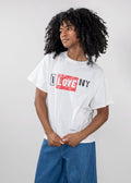 A person with curly black hair wearing a white graphic T-shirt with 'I LOVE NY' printed across the chest in black and red, paired with blue jeans, against a light gray studio background.