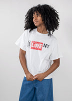 A person with curly black hair wearing a white graphic T-shirt with 'I LOVE NY' printed across the chest in black and red, paired with blue jeans, against a light gray studio background.