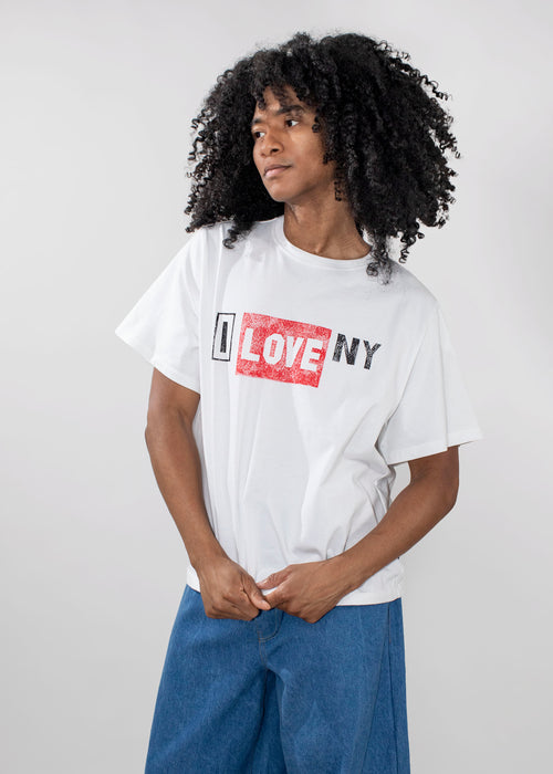 A person with curly black hair wearing a white graphic T-shirt with 'I LOVE NY' printed across the chest in black and red, paired with blue jeans, against a light gray studio background.