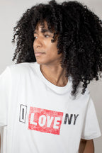 A person with voluminous black curly hair wearing a white short-sleeve T-shirt with a distressed red and black 'I LOVE NY' graphic on the front, photographed against a plain light gray background.