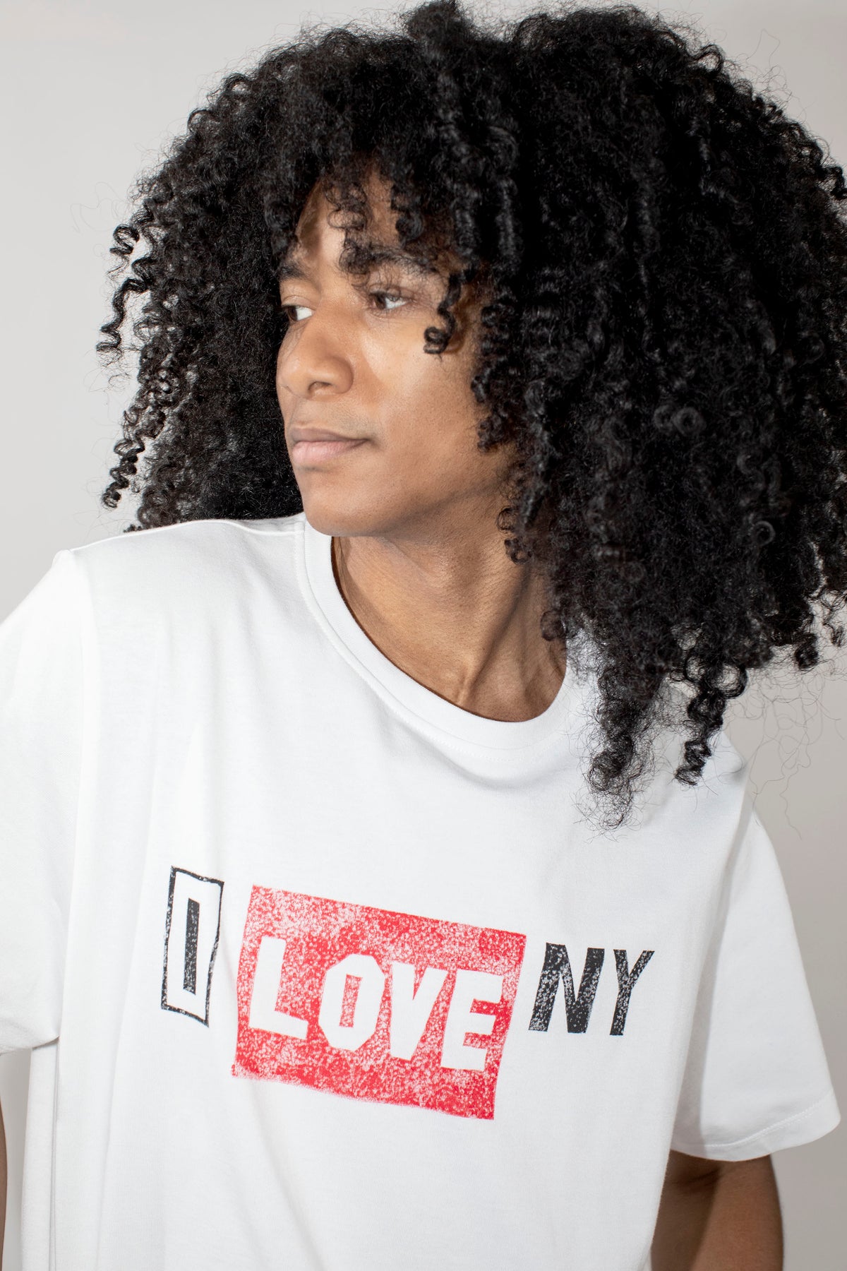 A person with voluminous black curly hair wearing a white short-sleeve T-shirt with a distressed red and black 'I LOVE NY' graphic on the front, photographed against a plain light gray background.