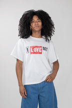 A model with curly black hair wearing a white graphic t-shirt with a red and black 'LOVE NY' print, paired with blue jeans, against a light gray studio background.