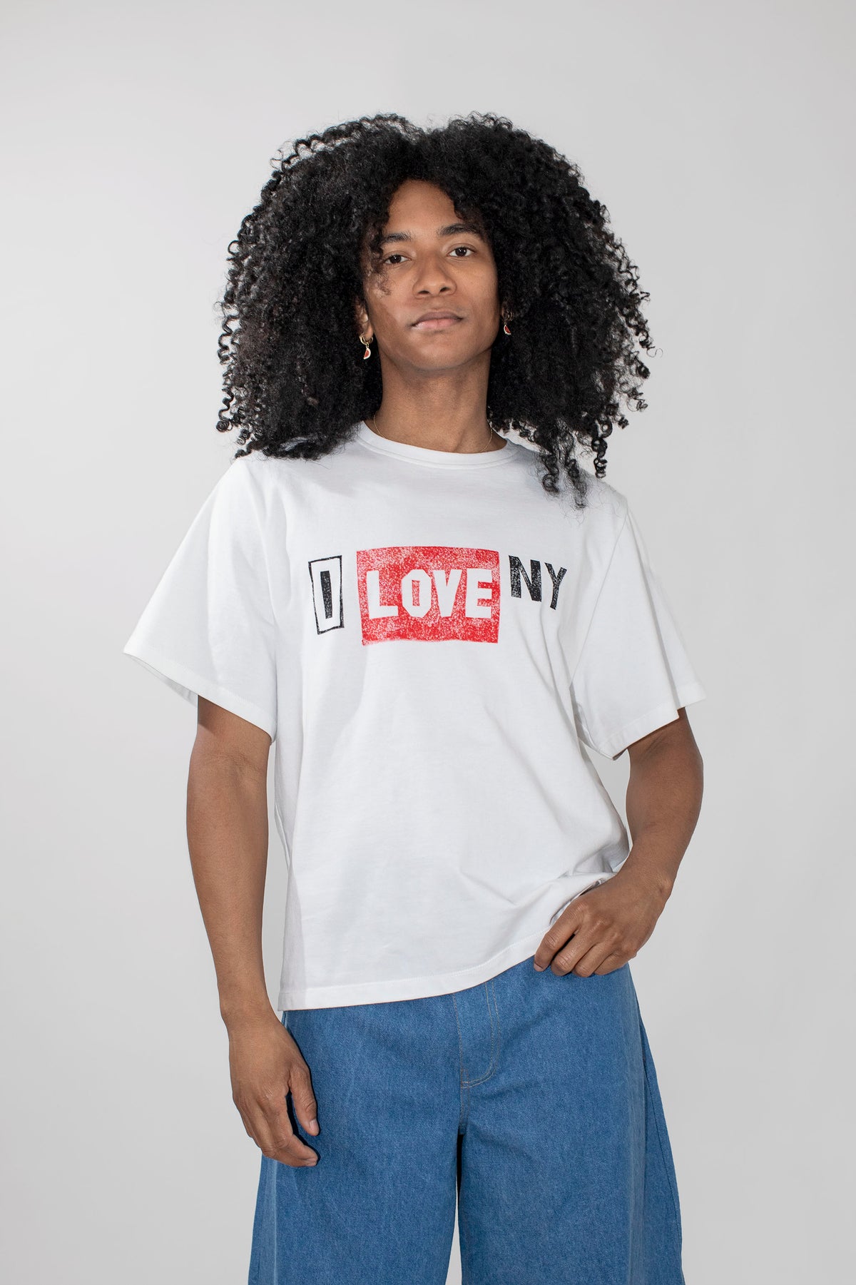 A model with curly black hair wearing a white graphic t-shirt with a red and black 'LOVE NY' print, paired with blue jeans, against a light gray studio background.