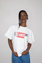 A model wearing a white short-sleeve t-shirt with a graphic print that reads 'I LOVE NY' in black and red, paired with blue jeans, against a plain light gray background.