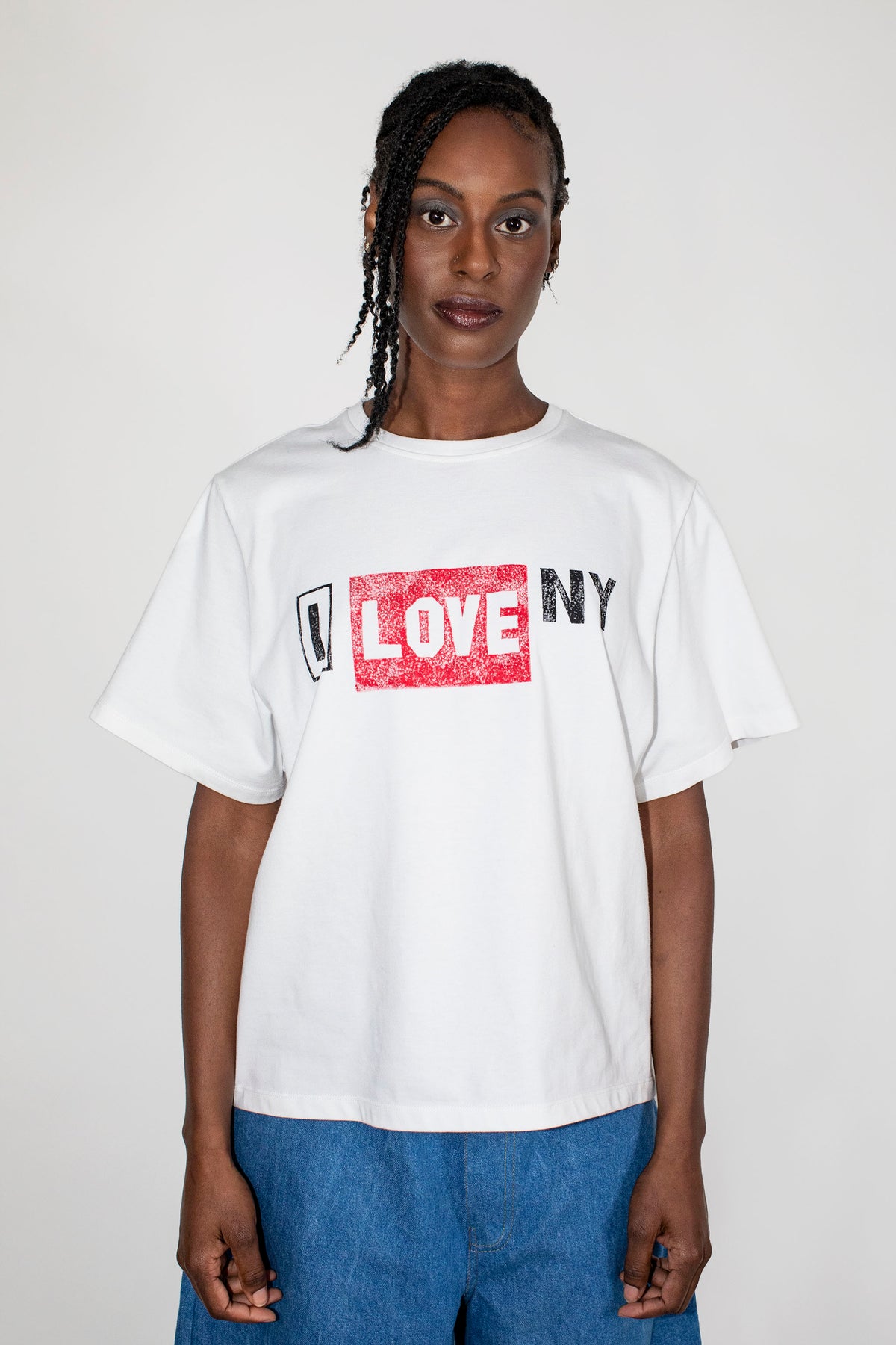 A model wearing a white short-sleeve graphic t-shirt with a distressed red and black 'I LOVE NY' print on the chest, paired with blue jeans, against a plain light gray background.