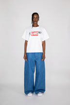 A model stands against a light gray studio background wearing a white graphic T-shirt with a red and black 'LOVE NY' box logo, paired with wide-leg blue jeans and white sneakers.