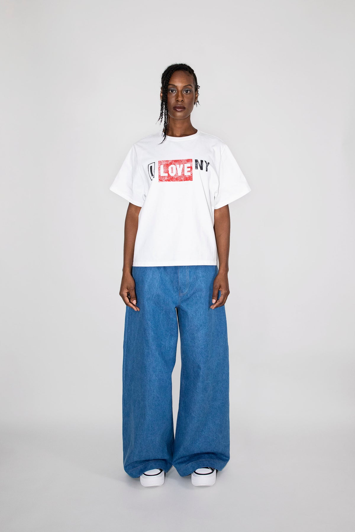 A model stands against a light gray studio background wearing a white graphic T-shirt with a red and black 'LOVE NY' box logo, paired with wide-leg blue jeans and white sneakers.