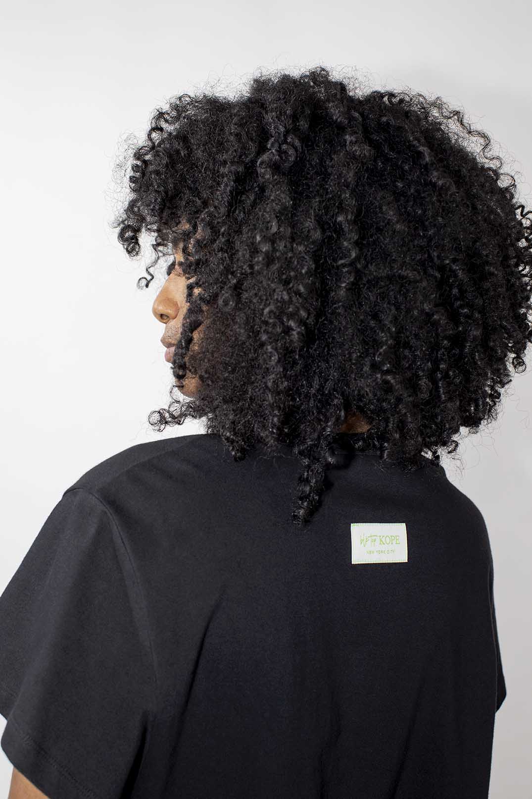 A person shown from the back with black short curly hair and a black T-shirt against a plain light gray background.