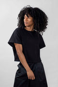 A model with voluminous curly black hair wearing a black short-sleeve t-shirt and black wide-leg trousers, standing against a light gray studio background.
