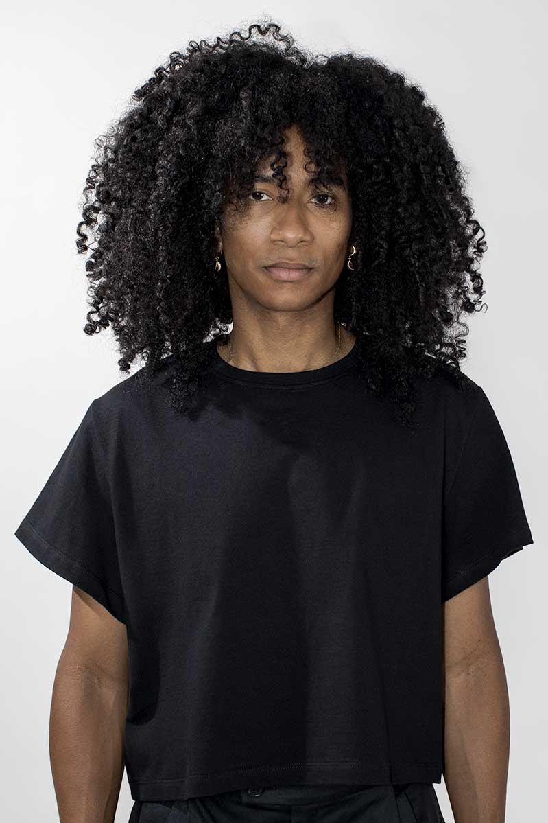 A person with voluminous curly black hair wearing a loose black short-sleeve T-shirt against a plain light gray studio background.