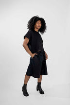 A model with curly black hair wearing a black short-sleeve t-shirt and matching black shorts, styled with black ankle boots, against a light gray studio background.
