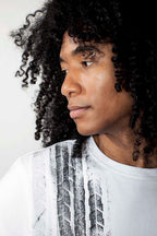 A close-up portrait of a person with curly black hair wearing a white crew-neck t-shirt with a black vertical tire-track style graphic print on the front, set against a plain light background.