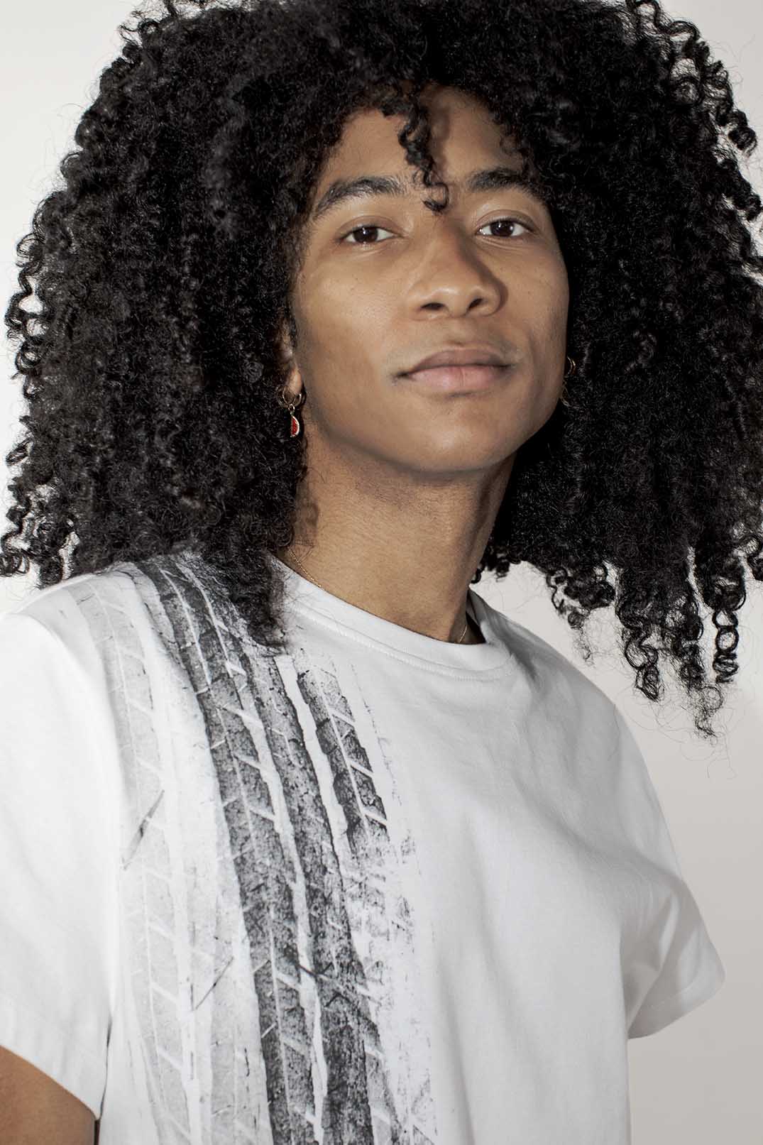 A close-up portrait of a person with voluminous dark curly hair wearing a white crew-neck t-shirt with a gray abstract vertical stripe pattern on the front, set against a plain light background.