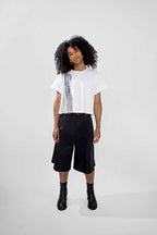 A model with curly black hair stands against a light gray studio background wearing a white short-sleeve T-shirt with a black vertical graphic print, black wide-leg cropped shorts, and black ankle boots.