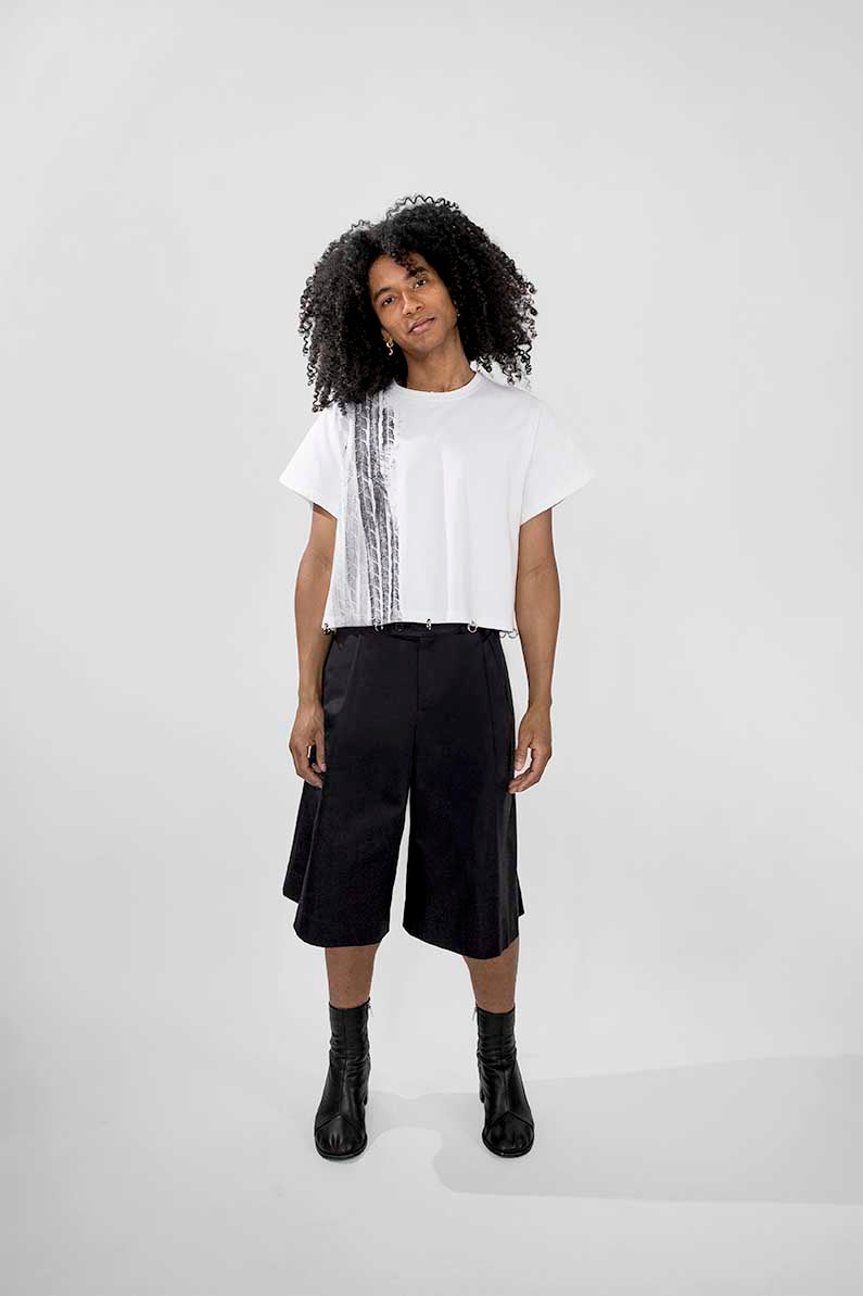A model with curly black hair stands against a light gray studio background wearing a white short-sleeve T-shirt with a black vertical graphic print, black wide-leg cropped shorts, and black ankle boots.