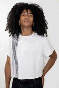 A model with voluminous curly black hair wearing a white short-sleeve t-shirt with a textured vertical black and white stripe detail on the left side, paired with black pants.