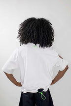 A person shown from the back wearing a white short-sleeve T-shirt and dark bottoms, with curly black hair, against a plain light gray studio background.