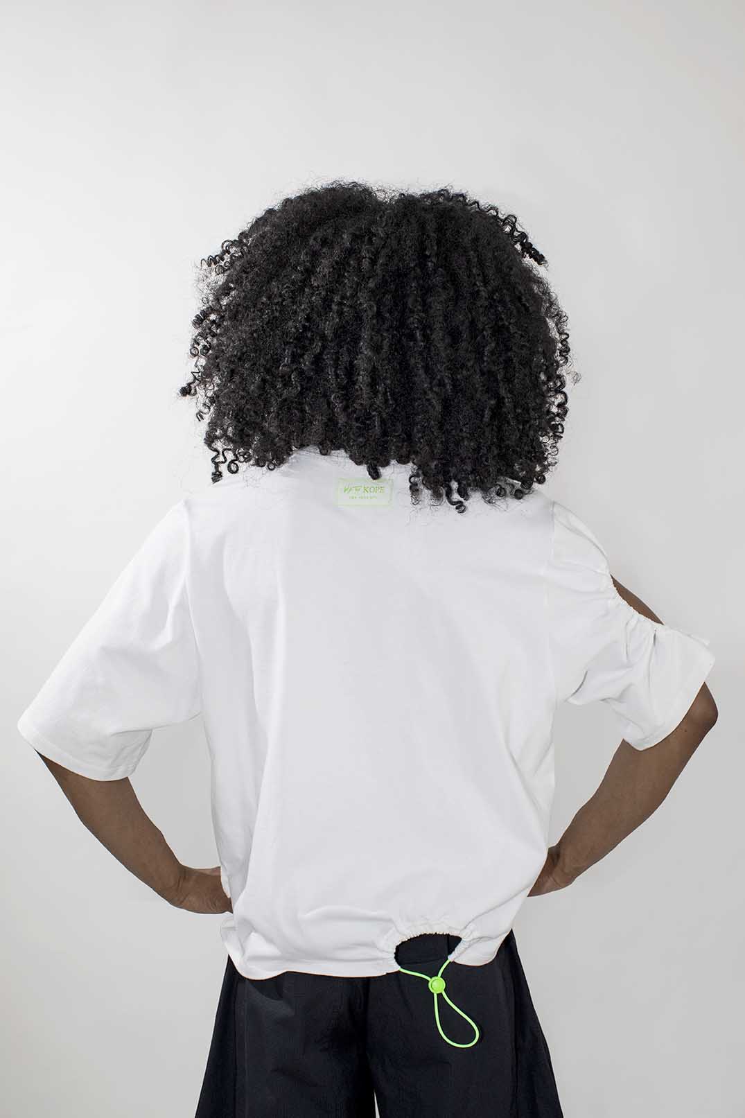 A person shown from the back wearing a white short-sleeve T-shirt and dark bottoms, with curly black hair, against a plain light gray studio background.