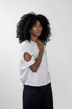 A model with curly black hair wearing a white short-sleeve top with neon green drawstring accents and black pants, standing against a light gray background.