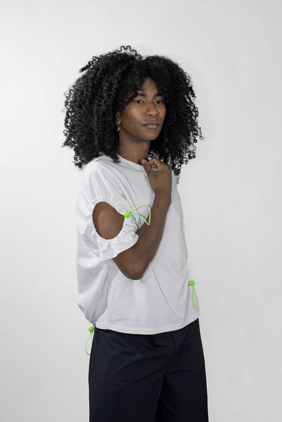 A model with curly black hair wearing a white short-sleeve top with neon green drawstring accents and black pants, standing against a light gray background.