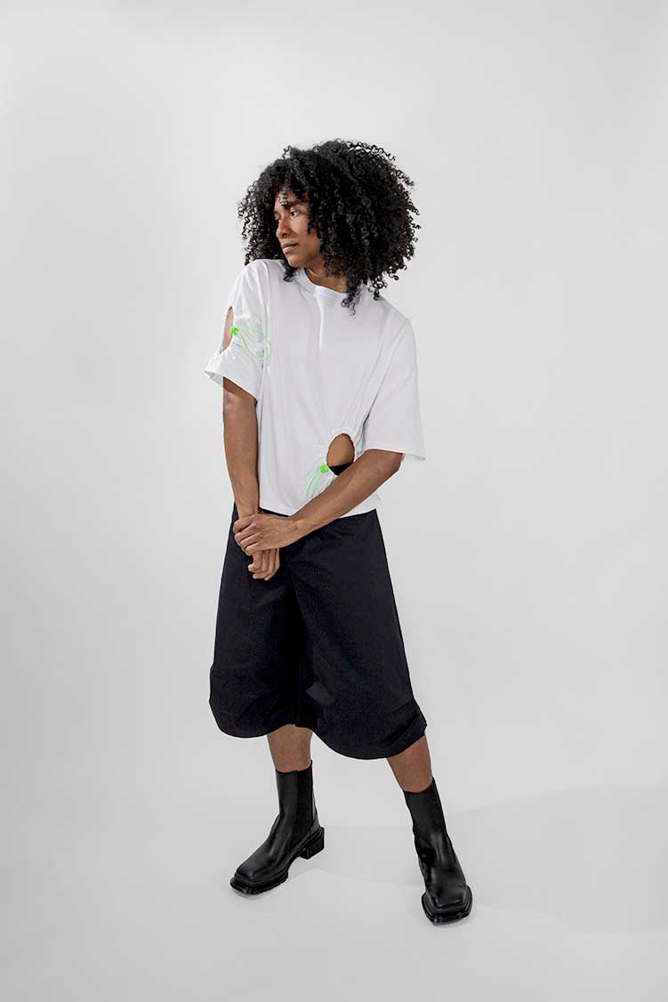 A model with curly black hair stands against a light gray studio background wearing a white short-sleeve top with cutout details, loose black shorts, and black ankle boots.