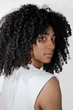 A person with voluminous, tightly curled black hair wearing a white sleeveless top, photographed against a plain light background.