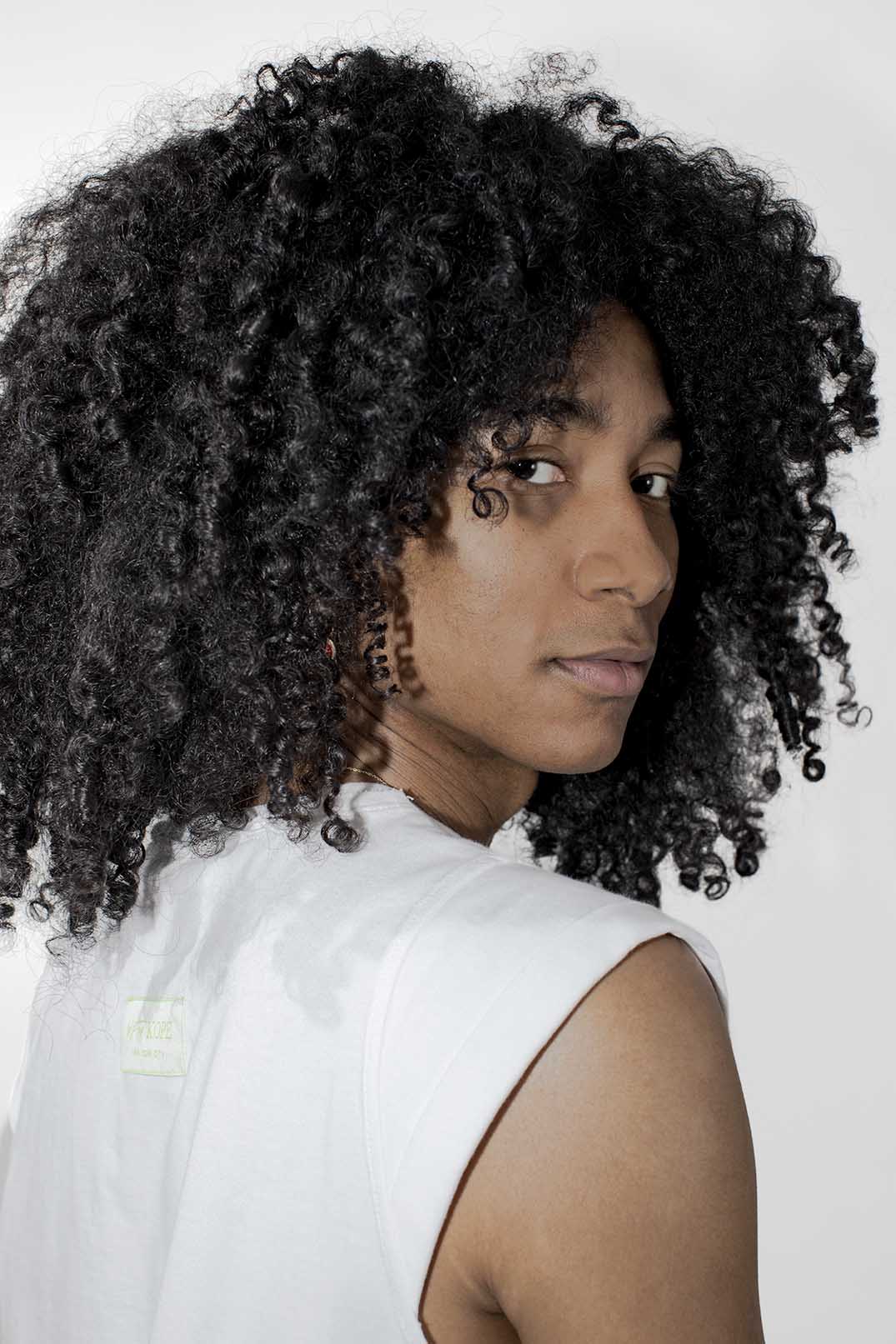 A person with voluminous, tightly curled black hair wearing a white sleeveless top, photographed against a plain light background.