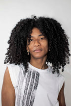 A person with voluminous, curly black hair wearing a white sleeveless t-shirt with a black graphic print, photographed against a plain light gray background.