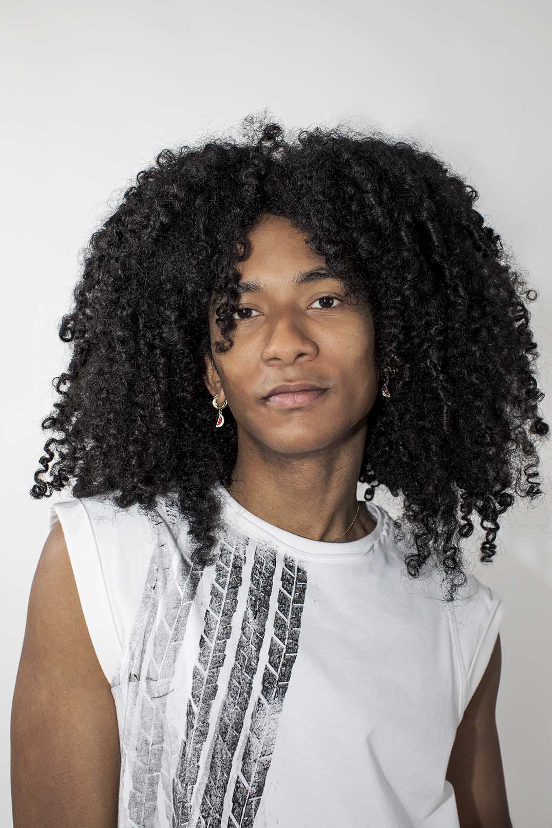 A person with voluminous, curly black hair wearing a white sleeveless t-shirt with a black graphic print, photographed against a plain light gray background.