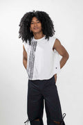 A model with long, curly black hair wearing a white sleeveless top with vertical black textured stripes and black wide-leg pants, standing against a light gray studio background.