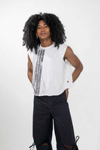 A model with long, curly black hair wearing a white sleeveless top with vertical black textured stripes and black wide-leg pants, standing against a light gray studio background.