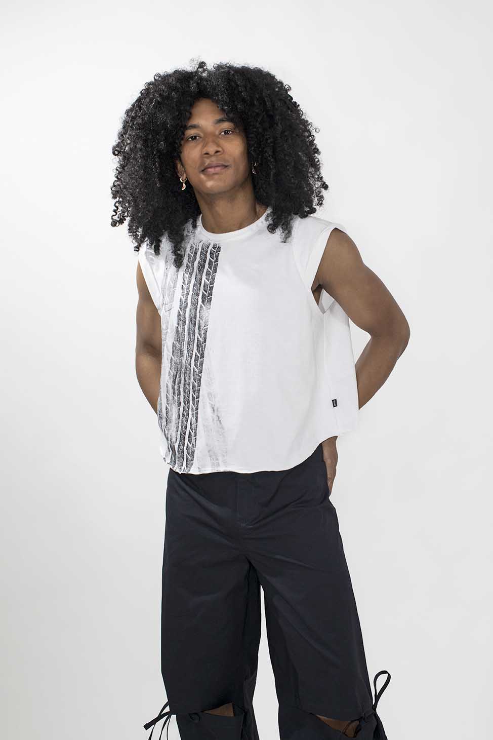 A model with long, curly black hair wearing a white sleeveless top with vertical black textured stripes and black wide-leg pants, standing against a light gray studio background.