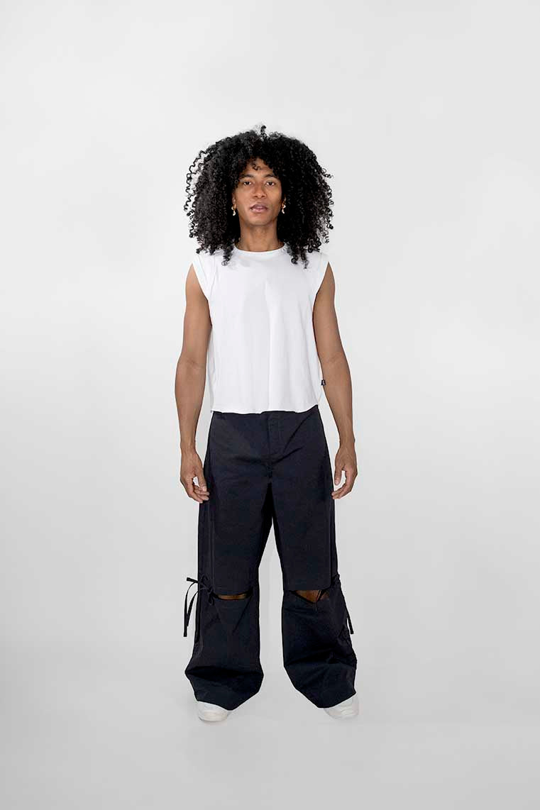 A model with curly black hair stands against a light gray studio background wearing a sleeveless white top and very wide-leg dark pants with knee cutouts and side drawstring details.