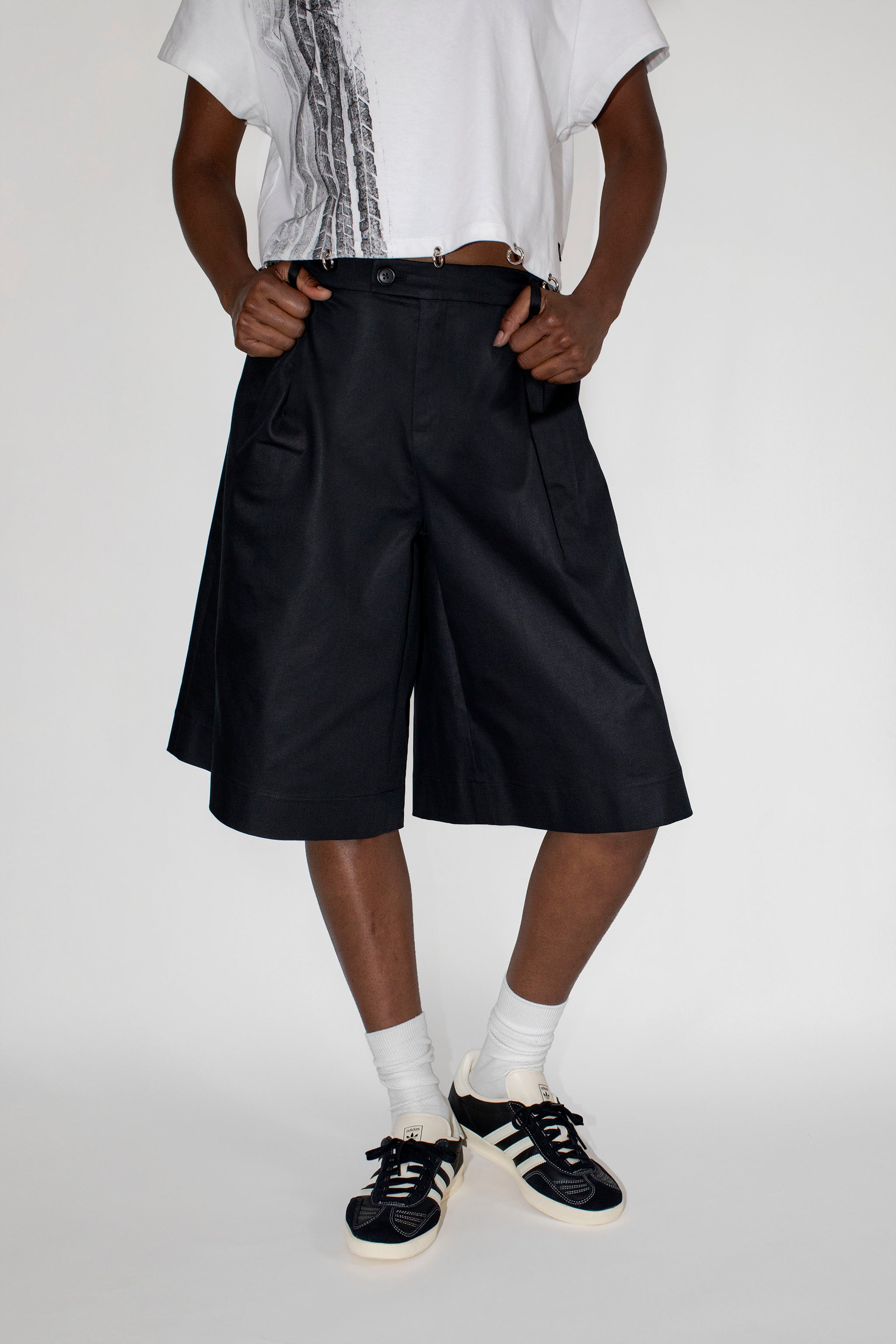 Person wearing black dress shorts, white shirt, and black and white sneakers on a plain background