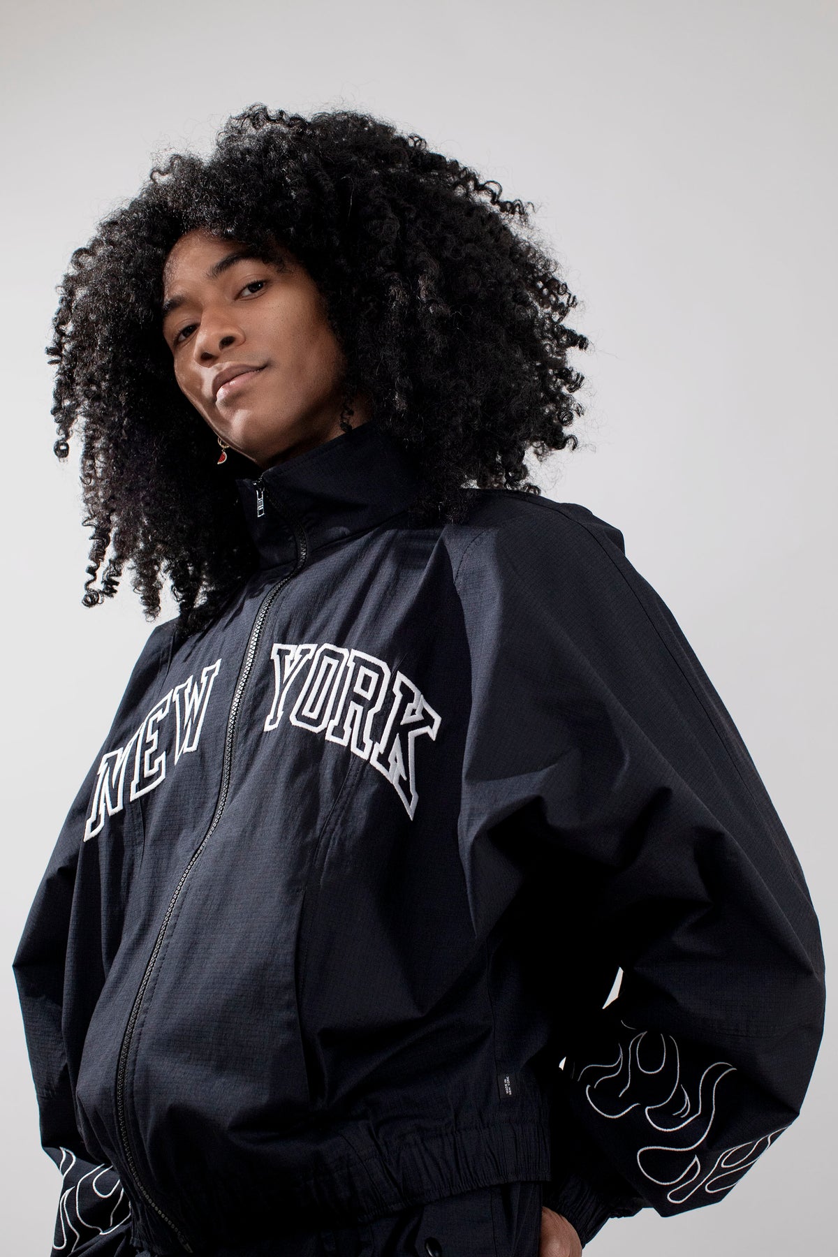 A person with voluminous dark curly hair wearing a black zip-up jacket with 'NEW YORK' embroidered in white arched letters across the chest and white flame-like embroidery on the sleeves, photographed against a plain light gray studio background.