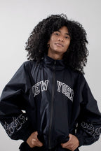A person with voluminous curly black hair wearing a black zip-up track jacket with 'NEW YORK' printed in white arched letters across the chest and white flame-like embroidery on the sleeves, photographed against a light gray studio background.