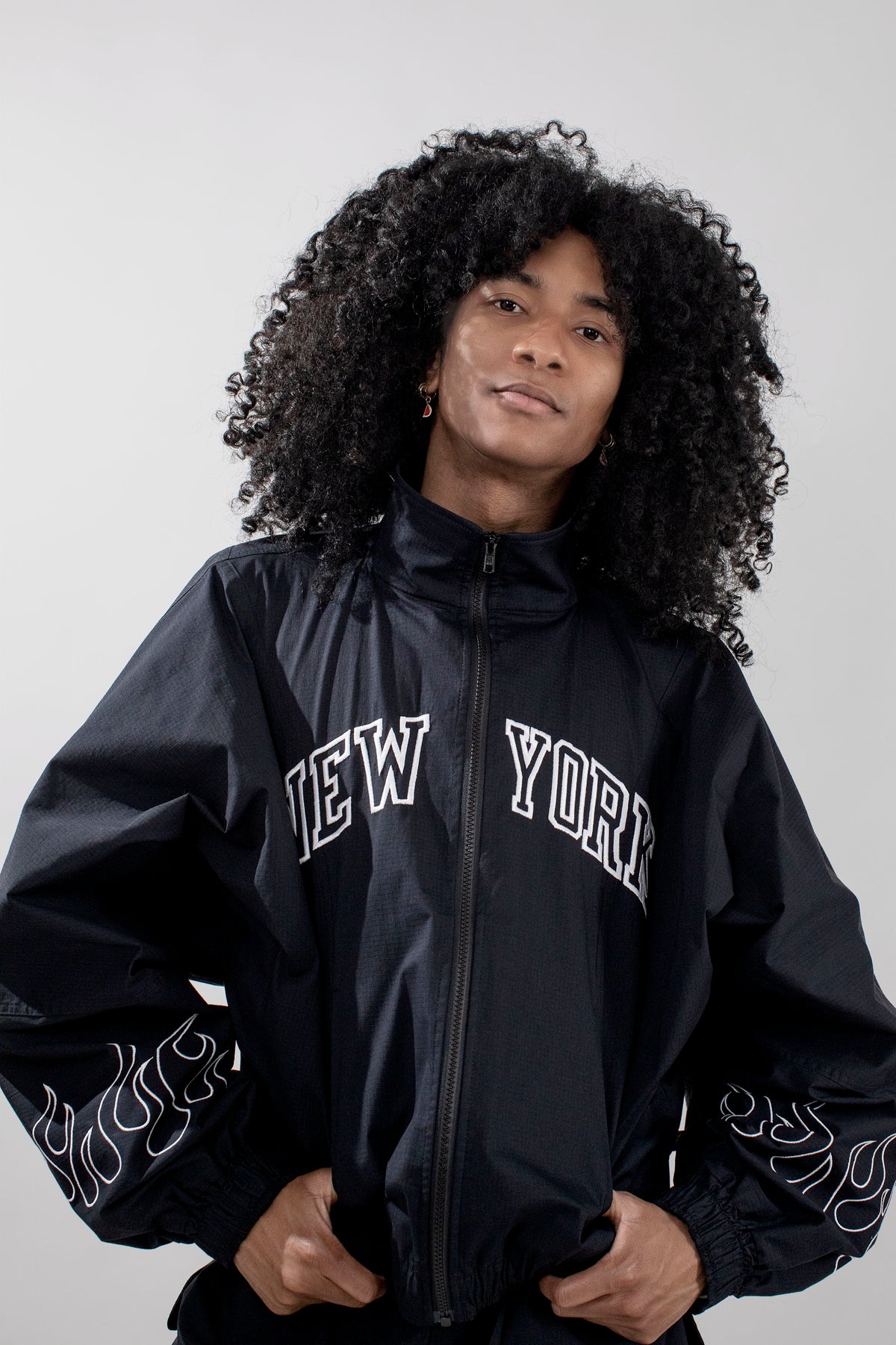 A person with voluminous curly black hair wearing a black zip-up track jacket with 'NEW YORK' printed in white arched letters across the chest and white flame-like embroidery on the sleeves, photographed against a light gray studio background.