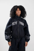 A model with curly black hair wearing a black zip-up jacket with 'NEW YORK' embroidered in white arched letters on the chest and white flame embroidery on the sleeves, paired with black pants, against a light gray studio background.