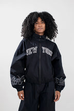A model with curly black hair wearing a black zip-up jacket with 'NEW YORK' embroidered in white arched letters on the chest and white flame embroidery on the sleeves, paired with black pants, against a light gray studio background.