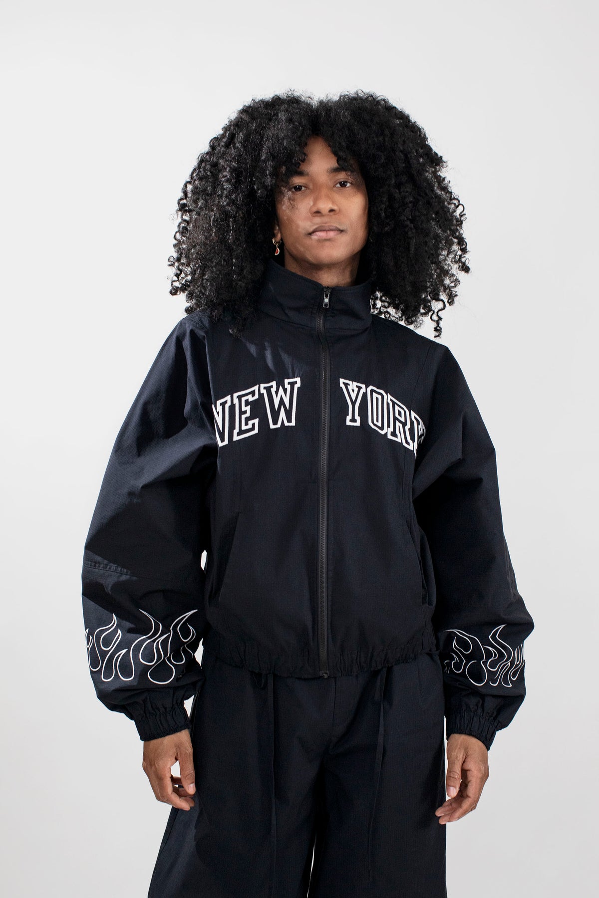 A model with curly black hair wearing a black zip-up jacket with 'NEW YORK' embroidered in white arched letters on the chest and white flame embroidery on the sleeves, paired with black pants, against a light gray studio background.