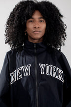 A person with voluminous curly black hair wearing a black zip-up jacket with 'NEW YORK' embroidered in white arched letters across the chest, photographed against a plain light gray background.