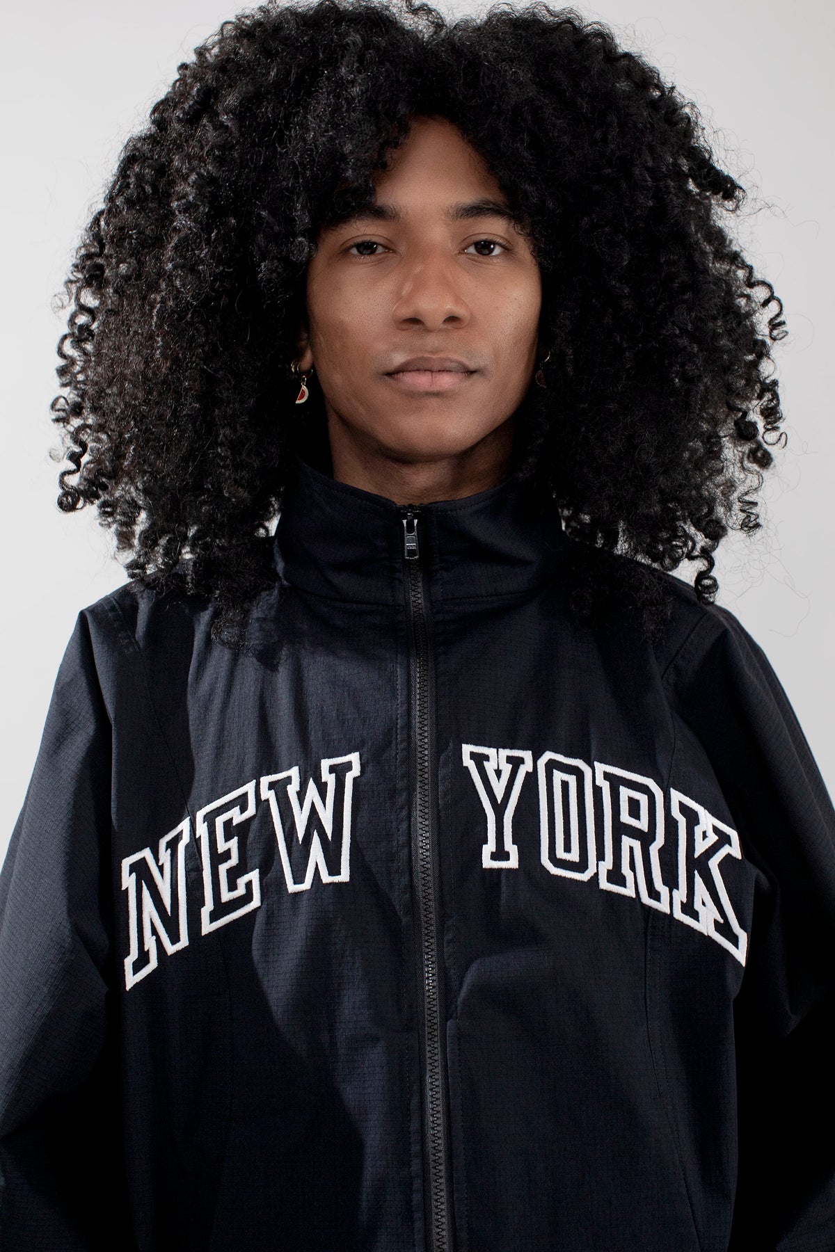 A person with voluminous curly black hair wearing a black zip-up jacket with 'NEW YORK' embroidered in white arched letters across the chest, photographed against a plain light gray background.