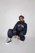 A model crouches in a studio setting wearing a black oversized zip-up track jacket with 'NEW YORK' embroidered in white across the chest, matching black jogger pants with white flame embroidery, and black-and-white high-top sneakers against a light gray background.