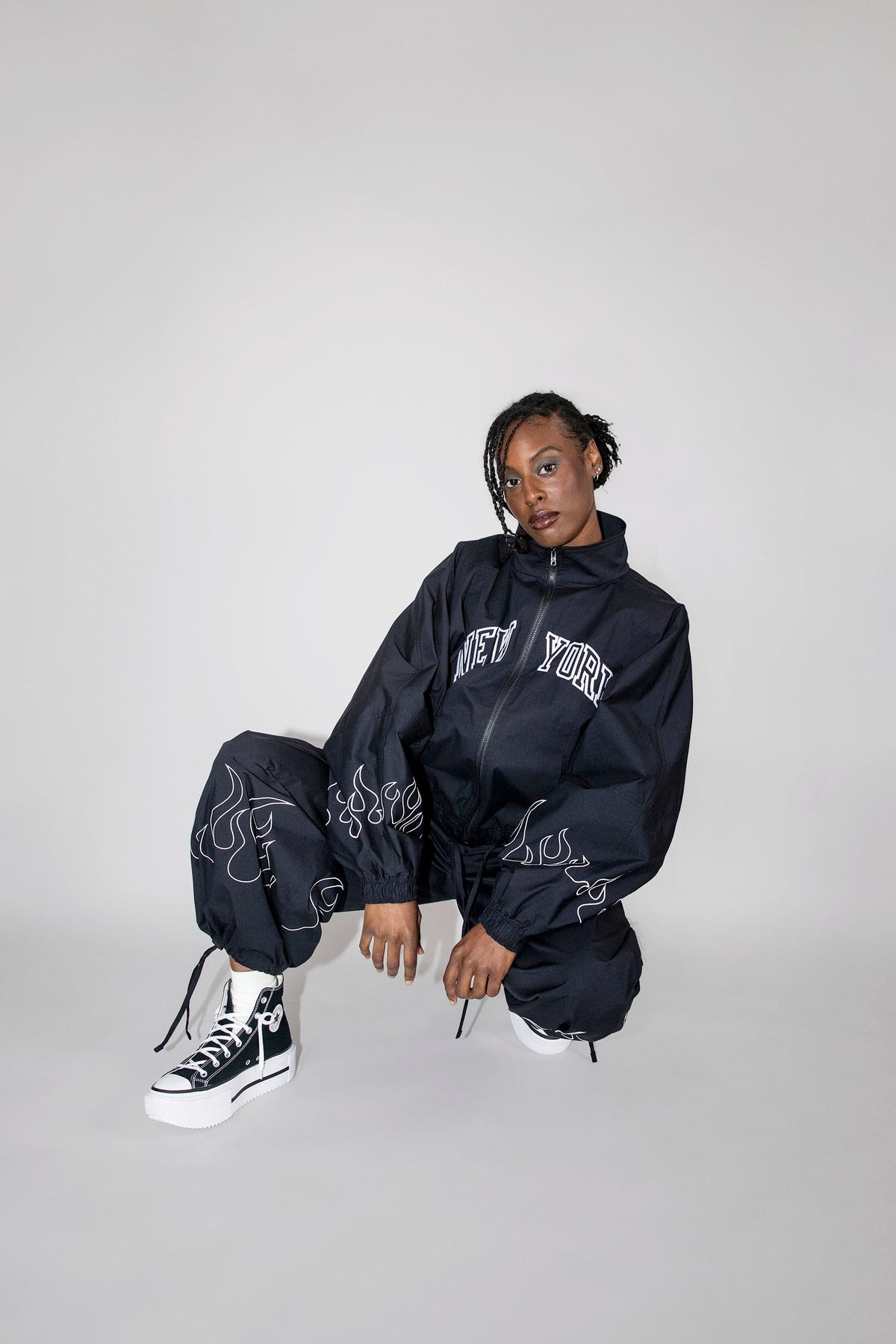 A model crouches in a studio setting wearing a black oversized zip-up track jacket with 'NEW YORK' embroidered in white across the chest, matching black jogger pants with white flame embroidery, and black-and-white high-top sneakers against a light gray background.