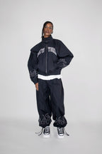 A model stands against a light gray studio background wearing a black zip-up jacket with 'NEW YORK' embroidered in white across the chest, matching black baggy pants with white flame embroidery near the hems, and white sneakers.