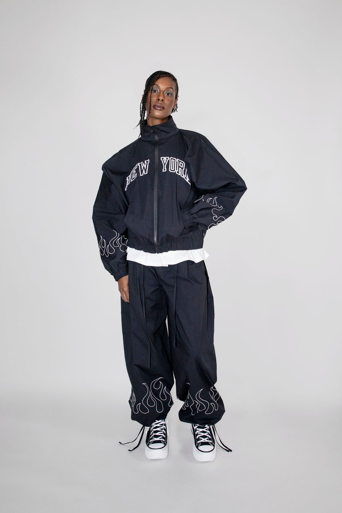 A model stands against a light gray studio background wearing a black zip-up jacket with 'NEW YORK' embroidered in white across the chest, matching black baggy pants with white flame embroidery near the hems, and white sneakers.