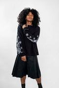 A model with curly black hair wearing a black long-sleeve top with white graphic patterns on the sleeves and a black pleated skort, standing against a light gray studio background.