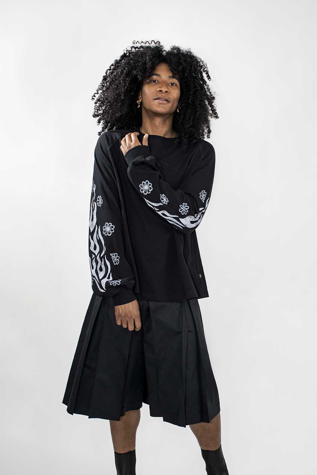 A model with curly black hair wearing a black long-sleeve top with white graphic patterns on the sleeves and a black pleated skort, standing against a light gray studio background.