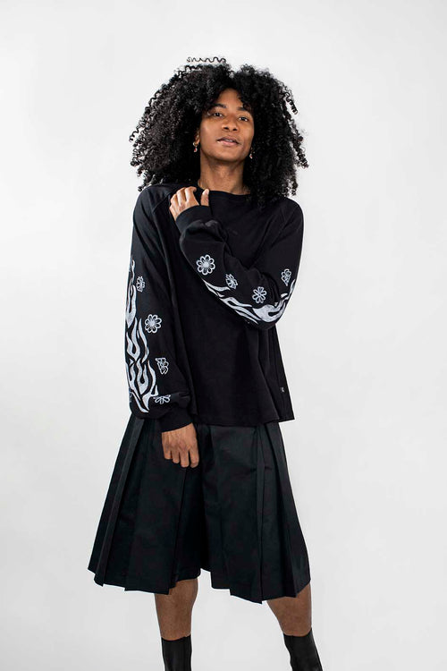 A model with curly black hair wearing a black long-sleeve top with white graphic patterns on the sleeves and a black pleated skort, standing against a light gray studio background.